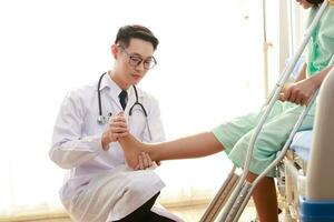 Asian male doctor examining the ankle of a female patient sitting on a nursing bed She holds a cane to help her walk. The concept of ankle pain, inflammation, injury. Physical therapy photo