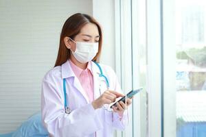 Beautiful Asian female doctor wearing a white mask stand holding a smartphone chat online through the application. Modern communication technology concept. Patient care through smartphones photo