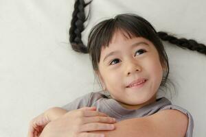 linda pequeño asiático niña con coletas ella sonrisas felizmente acostado en el blanco cama. positivo persona. foto