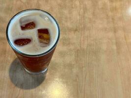 There is a glass of iced black tea covered with foam on the wood grain table photo