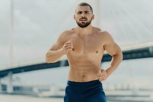 Serious male jogger runs actively, focused on the distance, posing with a naked torso against a blurred background with a bridge. Fit, muscular man trains actively. photo