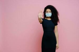 Ethnic woman with curly hair, making stop gesture, says no to COVID-19. Wearing mask, black dress, virus protection. photo