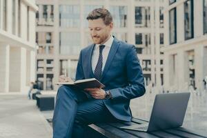 Relaxed office worker in blue suit, working outdoors on bench with laptop and agenda, city buildings blurred. photo