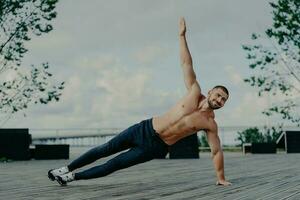 Fit man performs side plank, raises arm, trains outdoors, displays muscular physique, exudes satisfaction. Handsome athlete practices body balance, emphasizing sport. photo