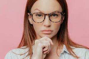 Confident business owner, chin in hand, raised eyebrow. Optical glasses, dark hair. Elegant shirt. Serious gaze. Studio shot photo