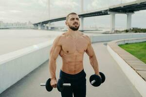 Concentrated bodybuilder with strong muscles holds barbells, exercises biceps near river bridge. Muscular, healthy lifestyle, sport concept. photo