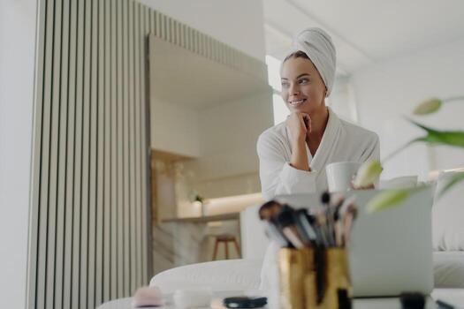 Beauty blogger in bathrobe with towel on head, smiles while relaxing on sofa with tea, manages blog on laptop. photo