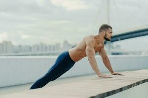Athlete in plank pose does push-ups outdoors, breathes fresh air. Strong body, naked torso. Motivated to stay fit and lead an active, healthy lifestyle. Sports training. photo