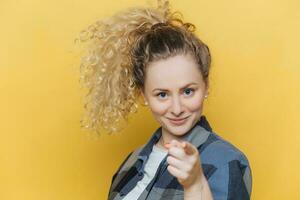 Confident woman chooses you, dressed casually, pointing at camera indoors. photo