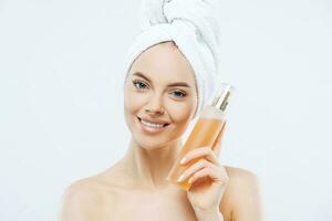 Smiling woman holds big floral perfume bottle, well-cared complexion, towel on head, shirtless photo