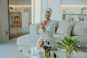 Pretty woman takes selfie, applies cream, cares for skin, in robe, towel, poses on sofa, does cosmetic procedures. photo