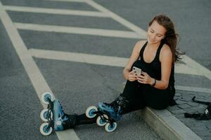 Top view of brunette active young woman uses smartphone for online communication poses on asphalt dressed in black active wear enjoys favorite hobby and free time. Extreme sport and rest concept photo