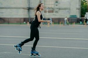 Rest leisure and lifestyle concept. Cheerful dark haired young European woman breathes fresh air while rollerskating has hobby leads active lifestyle poses outdoor engaged in fitness activity photo