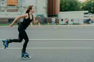 Horizontal shot of sporty energized woman enjoys rolleblading being photographed in motion poses on road against blurred street background has regular fitness activities for keeping fit and healthy photo