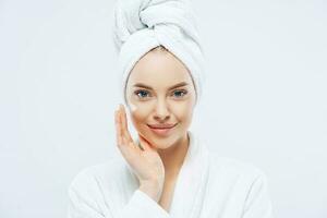 Young confident Caucasian woman applies face cream, enjoys new anti wrinkle cosmetic product, prevents sign of skin aging, wears minimal makeup, dressed in bath robe, isolated on white wall. photo
