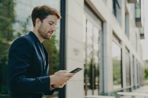 Focused man in dark suit chats, browses on phone near windowed building, typing message photo