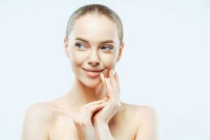 Lady gazes sensually, touches face, applies makeup, cares for skin, shirtless on white backdrop. Beauty, wellbeing concept photo