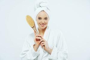 Pleased woman with healthy skin, gentle smile, holds body and foot massage brush, dressed in robe photo