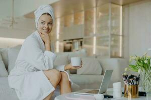 Cheerful woman in robe, fresh skin, drinks coffee, ponders, sits on sofa, works on laptop, uses cosmetics. photo