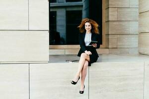 Confident young businesswoman in black suit and heels, holding gadget, exuding success, sitting crossed legs, camera-ready. photo