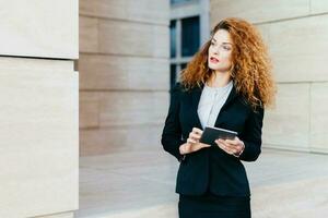 Curly-haired entrepreneur in formal attire plans work day, holds tablet, pensive gaze. Developing project with touchpad. photo