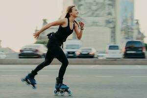 joven mujer sana delgada de cabello oscuro tiene tiempo de recreación disfruta patinar en velocidad se mueve rápido tiene expresión alegre usa camiseta negra y polainas. descanso activo y concepto de entrenamiento deportivo foto