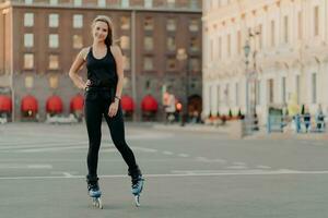 Sport and recreation concept. Horizontal shot of active woman practices rollerblading poses on blades keeps hand on waist blurred city background takes break. Rolleskating after work. Healthy life photo