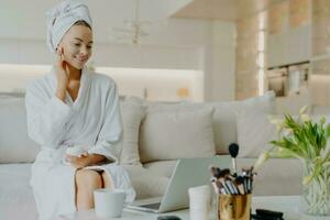 Smiling woman applies cream, advises skincare, in robe, towel, on sofa, rejuvenation procedure, laptop screen. Cosmetology. photo