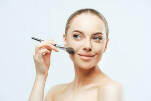 Facial treatment, cosmetology. Young woman, dark hair, applies powder with brush, sideways, naked body photo