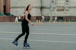 la mujer atlética en patines se mueve activamente posa sobre el asfalto en la calle vestida con ropa activa tiene una expresión alegre. joven modelo de fitness patinando en un lugar urbano. estilo de vida deportivo patinar foto