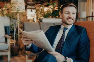 Seated entrepreneur reads newspaper, waiting for drink on cozy cafe sofa, in modern formal attire. photo