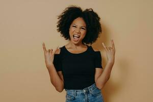 Young african woman with hands in rock n roll sign and shouting loudly, isolated on beige background photo