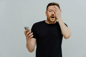 Scared man in black t-shirt covers half face, amazed at smartphone. Horrible video on display, reading social media post. Isolated on blue background. photo