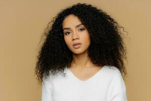 Serious dark-skinned female with frizzy hair, minimal makeup, calmly looks at camera. White jumper, brown background, deep in thoughts. photo