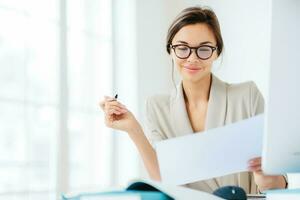 Concentrated businesswoman studies contract terms, writes with pen, dressed formally, at desk in spacious white interior photo