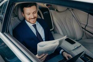 Relaxed entrepreneur in blue suit reads newspaper, checks news about successful company, works on notebook during commute. photo