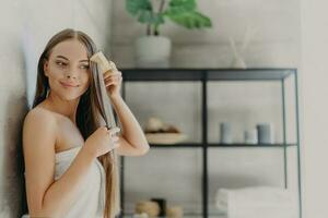 Smiling woman enjoys beauty, combs long hair, wrapped in towel in cozy bathroom, smooth skin. Hair care, spa concept. photo