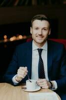 Satisfied employee in luxury suit, drinks coffee, smiles, sits at table, attractive look, awaits partner for important discussion. photo