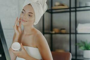 Pleased woman applies face cream, admires reflection, towel-wrapped, anti-aging after shower for healthy skin. Skin care concept. photo