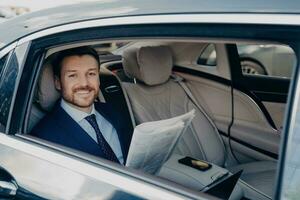 Smiling business owner in blue suit enjoys good news in newspaper, sitting in back of limousine during ride. photo