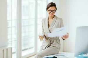 Pleased woman checks bills, dressed in beige suit, drinks, sits at desktop with monitor, holds chart sheet, poses by window photo