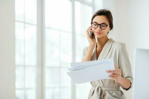 Business lady discusses contract details, works in office, focused on accounting information, holds documents during call, dressed formally photo