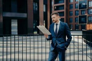 Handsome boss in blue suit stands thoughtfully, hand in pocket, reading newspaper for firm-related news, corporate buildings behind. photo