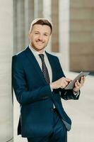 Vertical shot of business person uses digital tablet for online communication and work, uses free internet at office, has positive expression, dressed in formal black suit. People and occupation photo