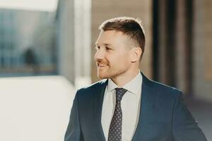 Sideways shot of cheerful unshaven male looks aside with joyful expression, wears formal clothes, being in good mood after successful meeting with business partners. People and work concept. photo