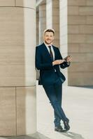 Photo of cheerful male dressed in expensive black suit, stands crossed legs, works on touch pad, stands in modern office interior, smiles positively at camera. Young male manager. Full length portrait