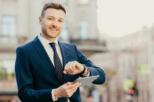 Shot of handsome male entrepreneur comes on meeting with business people, looks at wristwatch, uses touchpad for searching necessary information in internet, wears elegant formal suit and tie photo