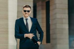 Positive glad young male wears formal suit, wears trendy shades, keeps hand in pocket, stands against office interior, being in good mood after successful business meeting. Leadership concept photo