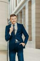 Thoughtful cheerful bearded male entrepreneur has pleasant smile, wears black suit, holds sunglasses, poses indoor, being delighted with successful business meeting and signing contract with partner photo