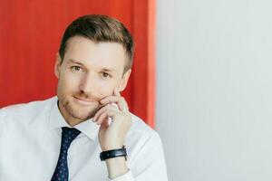 Confident office worker in white shirt with tie, hand on cheek, contemplates new stage in life. Poses against red and white background. Business concept. photo